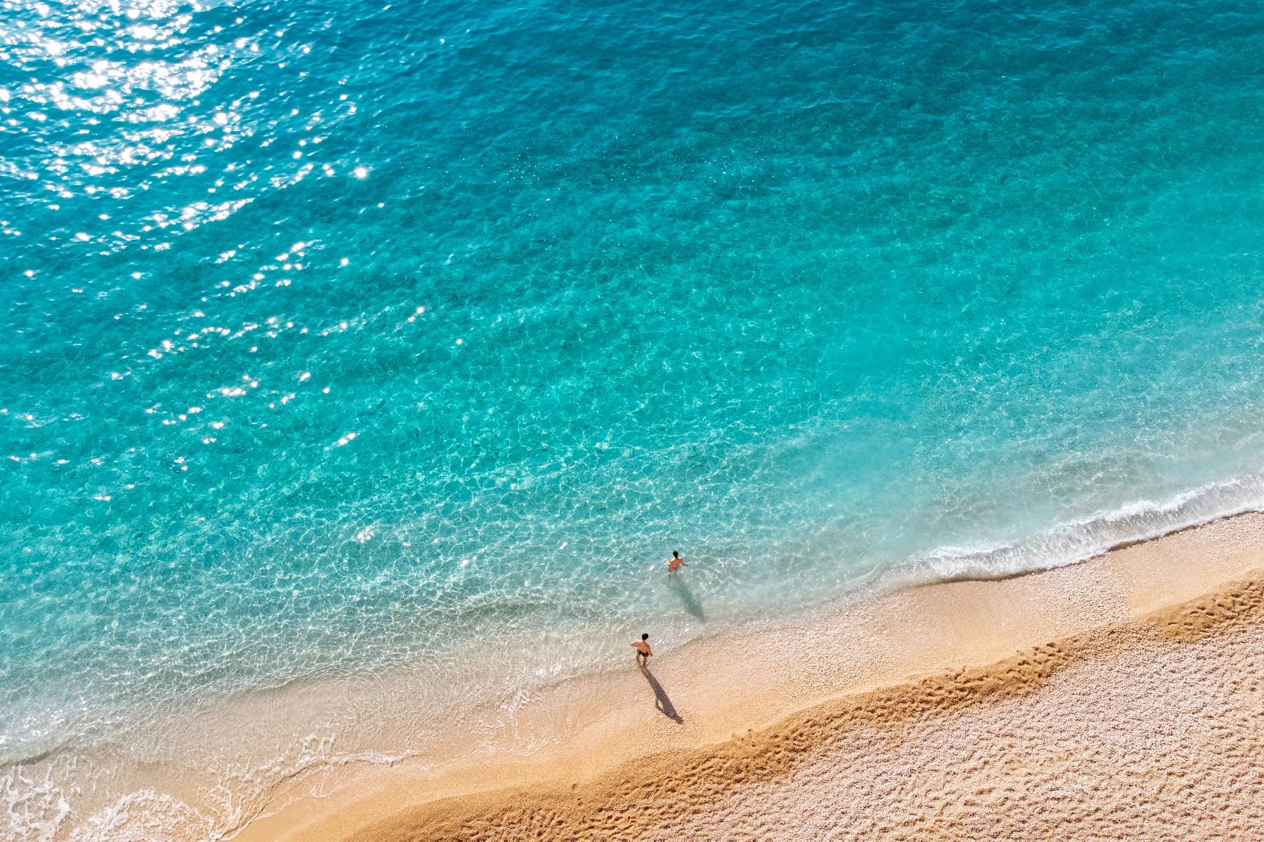 Two people at the edge of turquoise water on a sand beach, seen from above